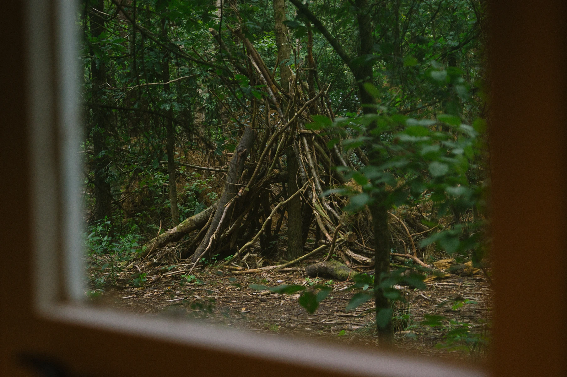 Photo of a hut build with branches looking out at it from a window in a cabin.